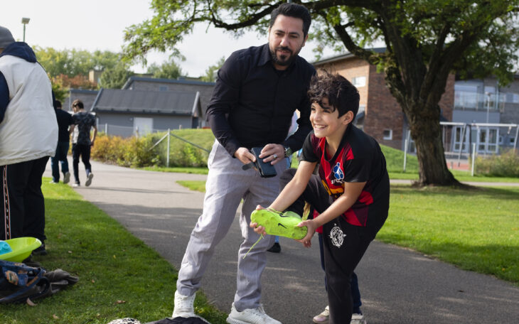 En leende pojke provar en fotbollssko, medan hans pappa står med mobilen i handen.