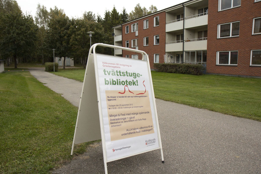 På bilden syns en hyreshuslänga i tegel med tre våningar i bakgrunden. I förgrunden står en så kallad gatupratare på vilken det står "Välkommen till invigning av Gråstensgatans tvättstugebibliotek", tillsammans med datum och tid. Undertecknat av Hyresgästföreningen och Hudiksvalls Bostäder. Gatuprataren står till hälften på en gräsmatta, till hälften på en asfalterad gång- och cykelväg.