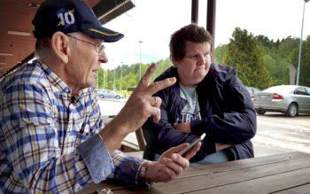 Ove Fafeeng och Andreas sitter vid en bänk. I bakgrunden ses bilar på en parkering.