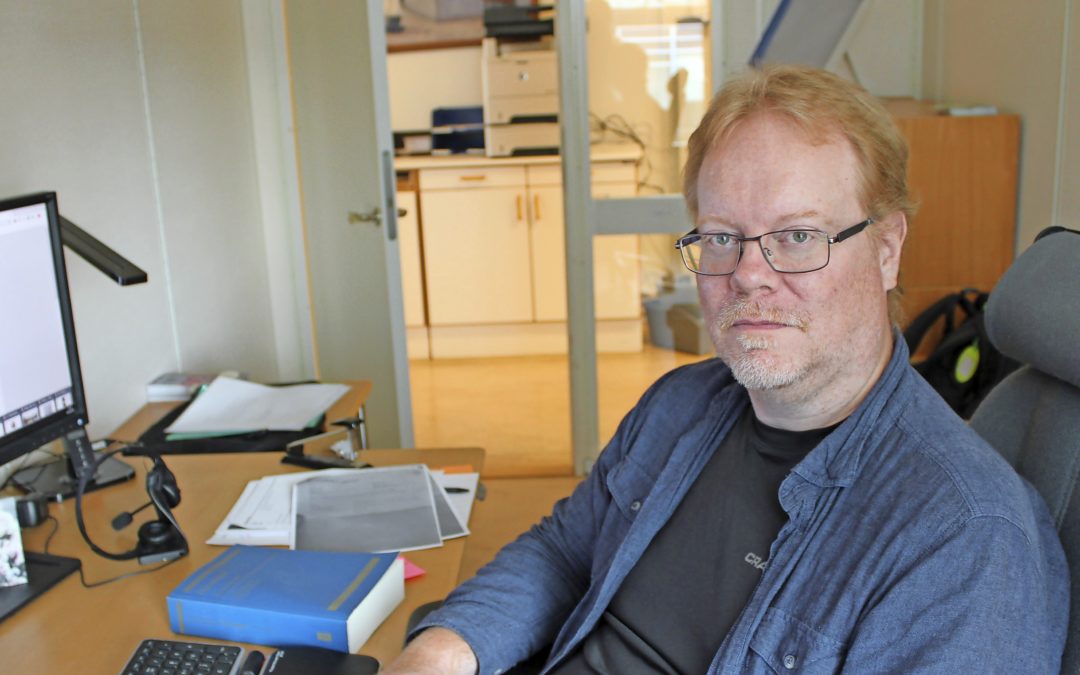 En man med rött hår och glasögon, blå uppknäppt skjorta med svart t-shirt under, sitter vid ett skrivbord. Framför sig har han ett tangentbord och bredvid lagboken. Bilden är i halvfigur.