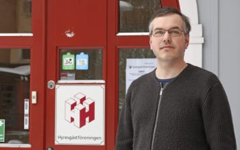 Marcus Kjellin i en brun tröja med blixtlås. Han står utanför en röd dörr där Hyresgästföreningens logga syns