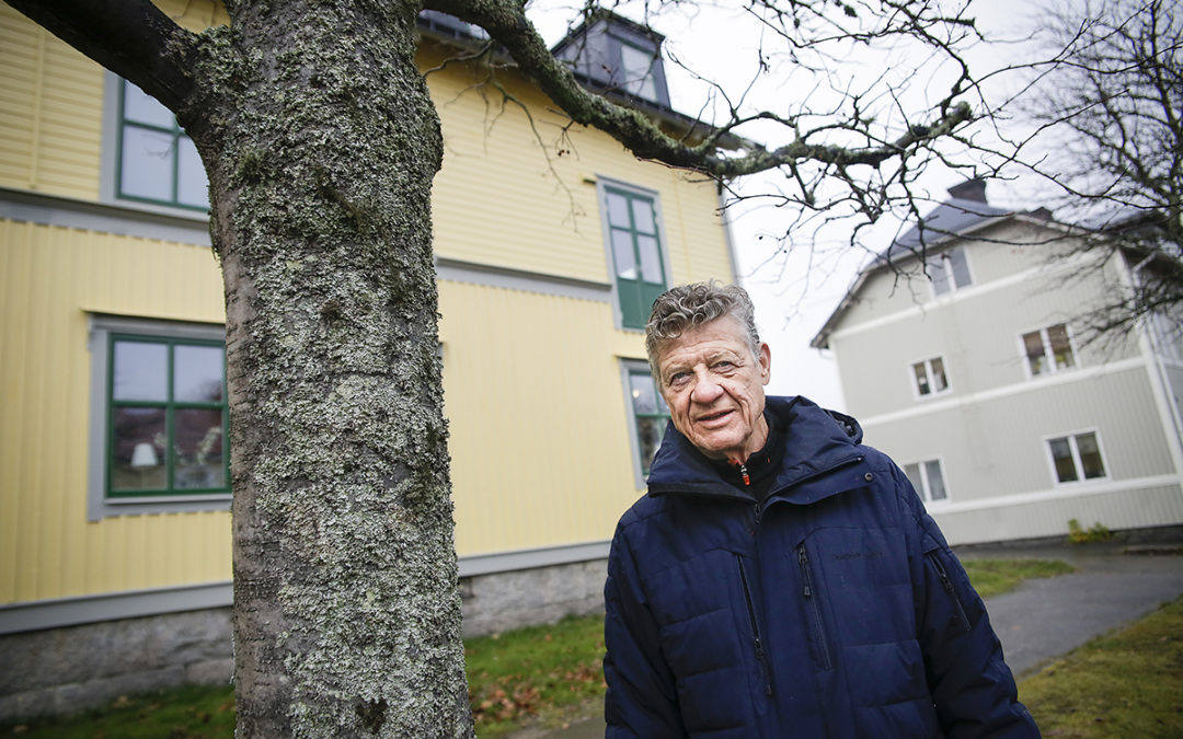 Foto på Greger Persson i mörkblå vinterjacka som står bredvid ett träd. I bakgrunden syns ett gult och ett ljusgrått trähus.