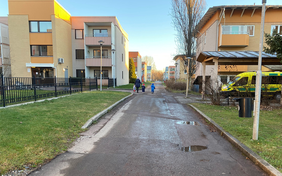 Bilden visar en gångväg som kantas av hus i tre våningar. En bit bort står en sjuktransport-bil och ännu längre bort ser man en kvinna och två små barn. Himlen är blå men det ligger vattenpölar på marken. Det är höst ute.