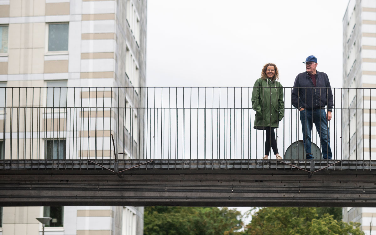 Monica Nilsson och Christer Jönsson, står på en bro i området Almgården i Malmö. Vita hus ramar in bron.