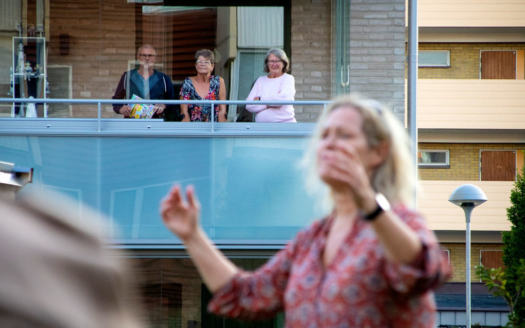 En kvinnligt dirigent dirigerar. Hon står utomhus framför en balkong. På balkongen står tre hyresgäster.