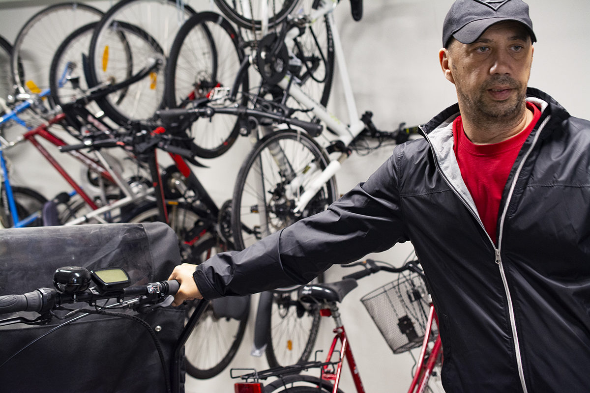 Samir Temic står inne i cykelrummet med en lådcykel från cykelpoolen. På väggarna hänger vanliga cyklar som tillhär hyresgästerna upphängda. 