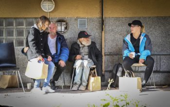 En kvinna sitter i knät på en man. Bredvid dem sitter en man som tittar bort mot en kvinna i keps. Alla fyra sitter på stolar mot en vägg.