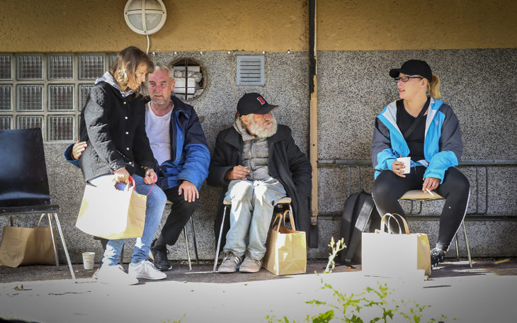 En kvinna sitter i knät på en man. Bredvid dem sitter en man som tittar bort mot en kvinna i keps. Alla fyra sitter på stolar mot en vägg.