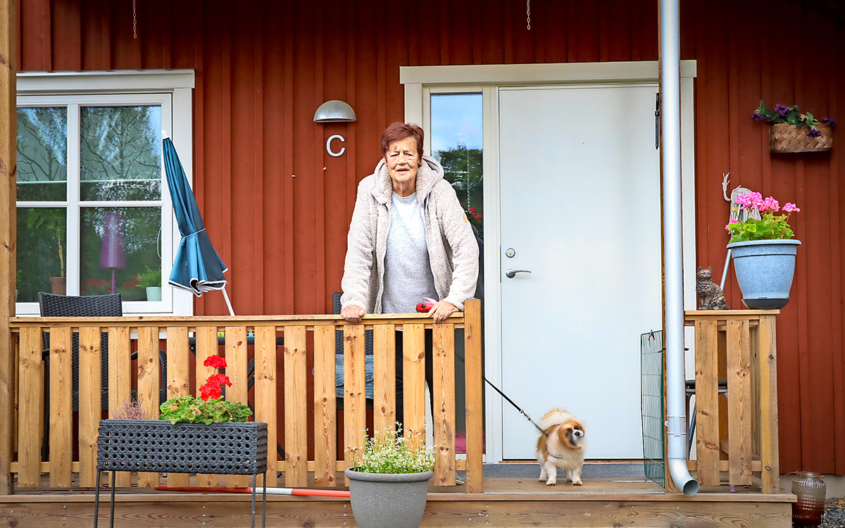 En kvinna som står på den lilla verandan utanför sin bostad. Hon lutar sig mot ett räcke och bredvid henne syns hennes lilla hund.
