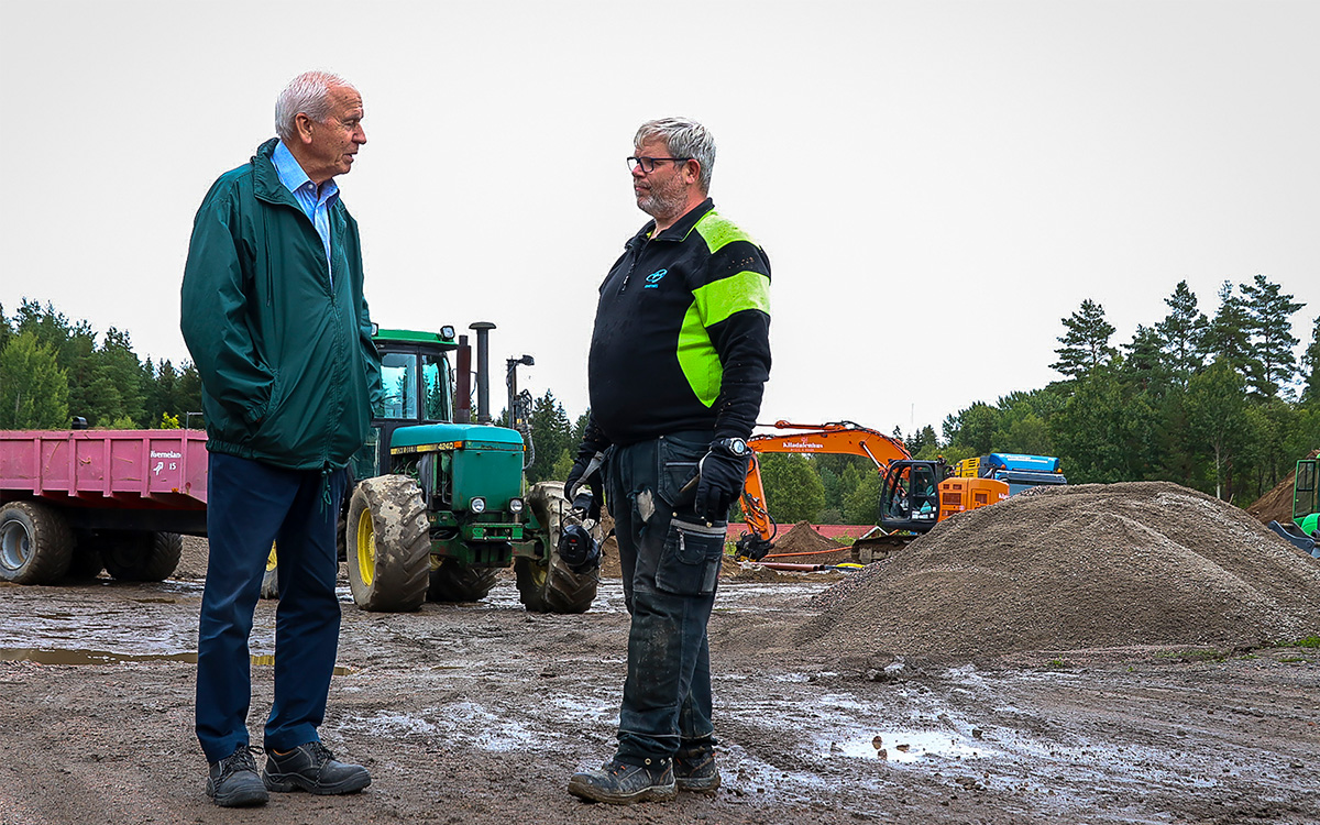 Två män står och talar med varandra på en lerig byggplats. I bakgrunden syns en traktor. Här ska byggas fler hyreshus på landet.