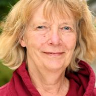 Bilden visar ansiktet av en kvinna i 50-60-årsåldern. Hon har rödlätt hår som är klippt i en pagefrisyr med lugg. Hon ler svagt och tittar in i kameran. Hon har en röd tröja med krage eller huva som sticker fram.