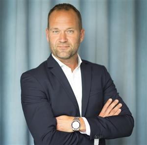 Bilden visar en man som står med armarna i kors ch tittar rakt in i kameran. Han har kortklippt mörkt hår som tunnas ut vid tinningarna och lite skäggstubb. Han är klädd i en vit skjorta och mörkblå kavaj. Han syns i halvfigur mot ett draperi som är svagt turkosfärgat. På vänstra handleden har han en klocka.