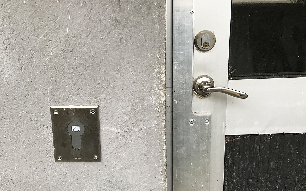 Till vänster i bild ett elektroniskt lås, en metallbricka med en stor nyckelhålsformad öppning som man ska lägga sin låstagg mot och i den Gavlegårdarnas logga. Brickan sitter på en grå, lite smutsig och liten vägg. Till höger ett dörrhandtag och ett ovanför det et lås som ser ut att gå att öppna med vanlig nyckel. Dörren, som man bara ser en del av, är i ljus metall med en glasruta upptill och en svart ruta nertill. Sidan av dörren är färstäkt med en metallkarm som ska skydda mot inbrott.