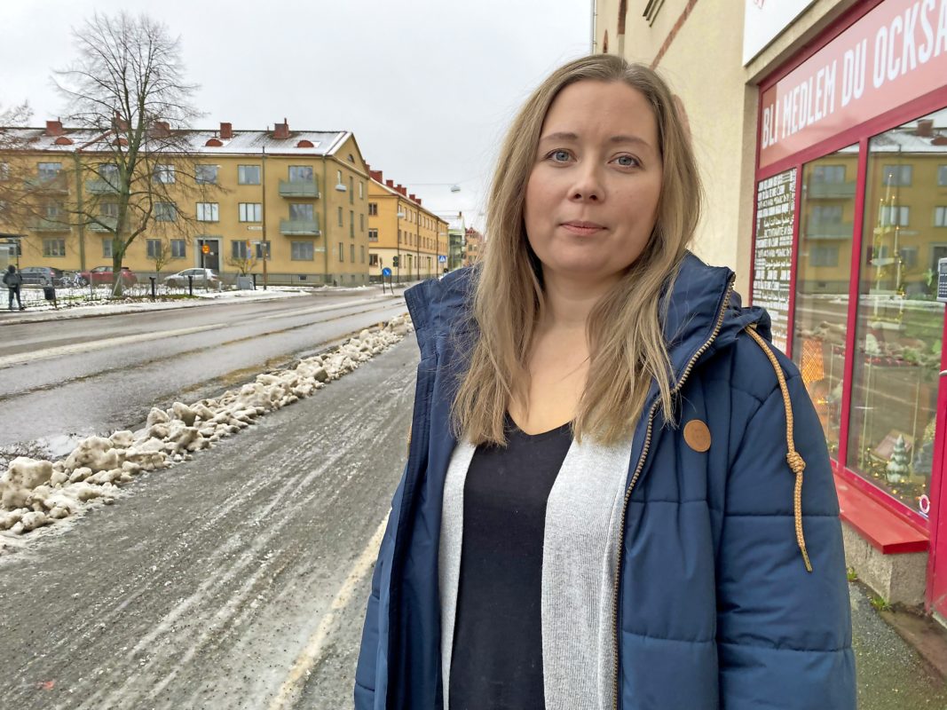 Kvinna i blå jacka står på en gata med snöslask framför ett stenhus.