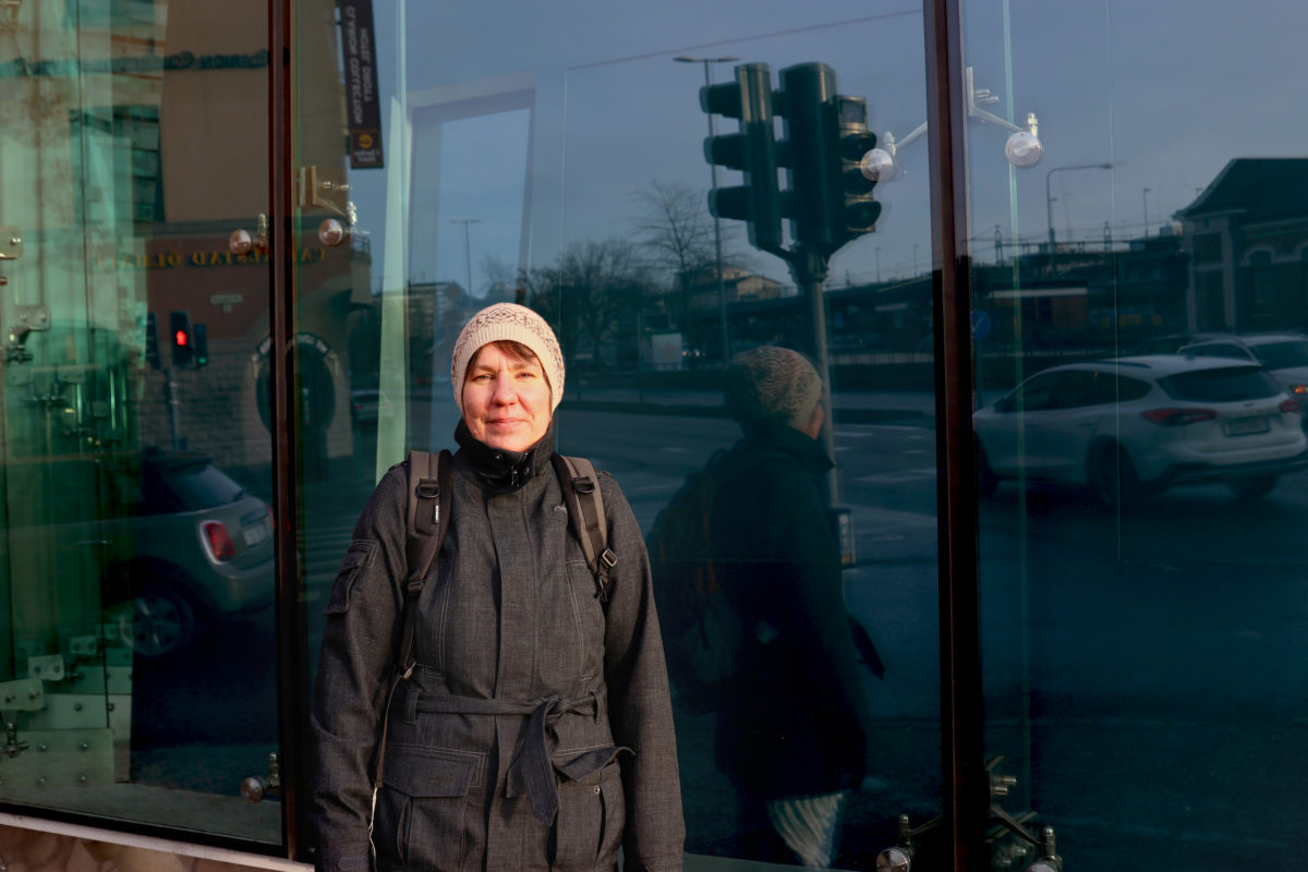 Kvinna med mössa som ser in i kameran står framför en glasvägg där trafikljus och bilar reflekteras.