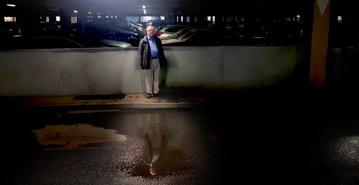 En man står i ett garage och hans spegelbild reflekteras i en vattenpöl.