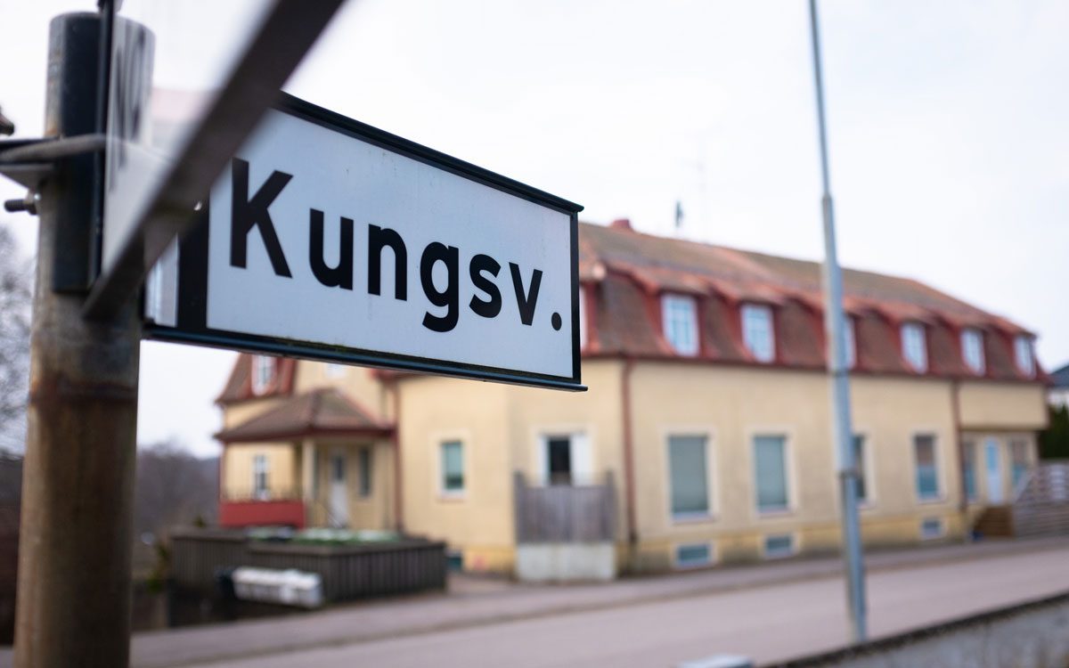 En skylt med texten "Kungsv." och ett gult bostadshus i bakgrunden.