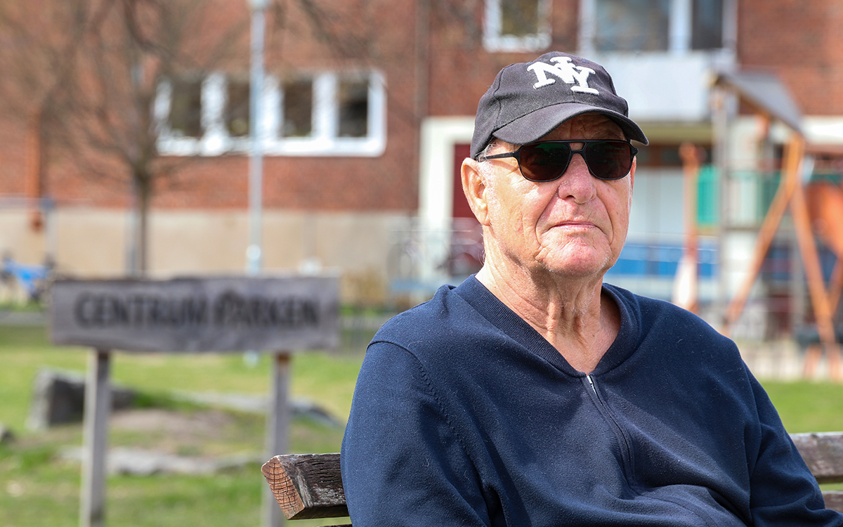 Man i keps och mörka solglasögon. Han sitter på en parkbänk. I bakgrunden syns en gräsmatta och delar av ett tegelhus.