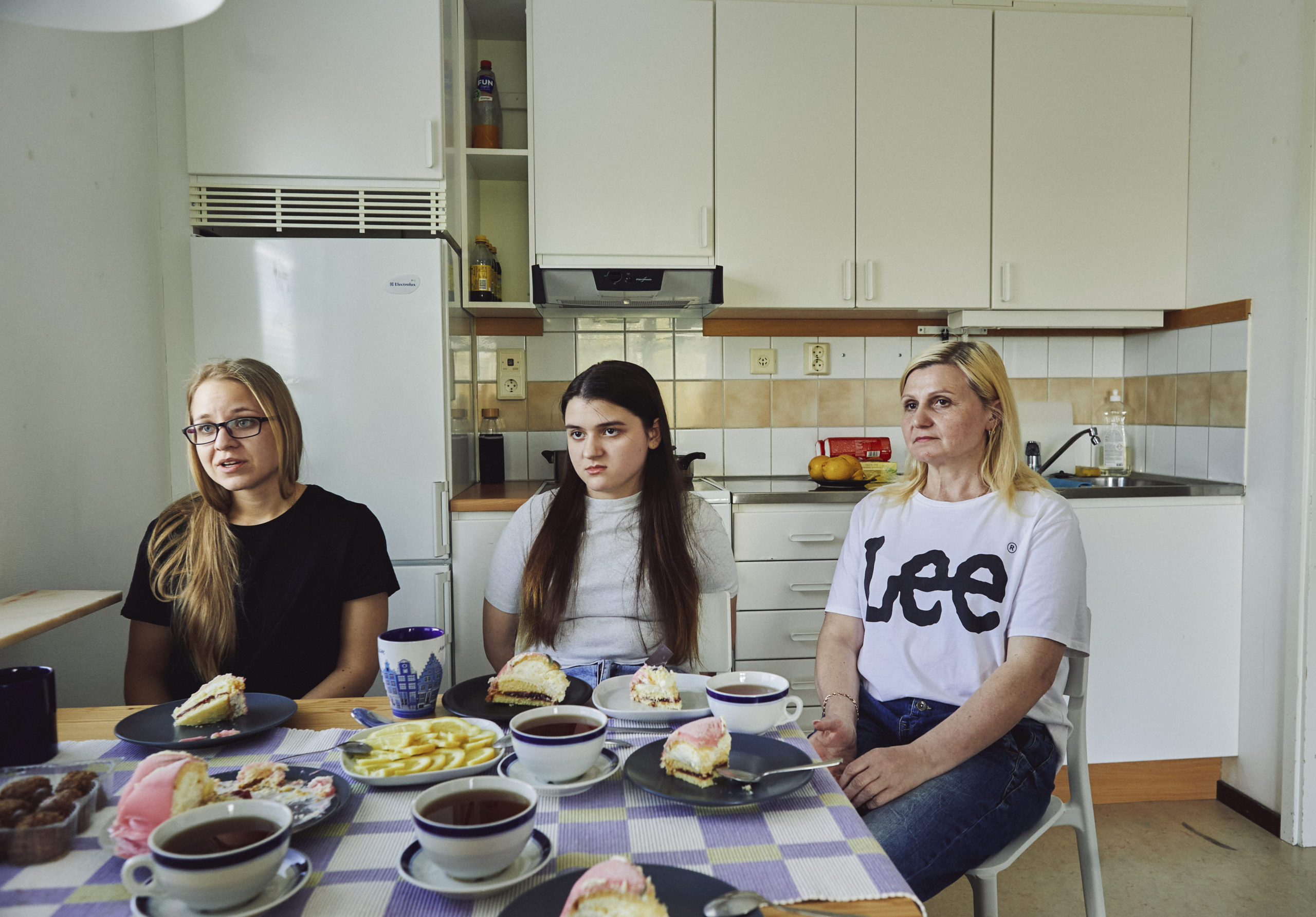 Tre personer, två kvinnor och en flicka, sitter vid ett köksbord. De har allvarliga miner. Kvinnan längst till vänster är Viktoria 29 år. Hon har långt blont hår, svarta rektangulära glasögon och har på sig en svart t-shirt. Bredvid henne sitter 14-åriga Viola. Hon har långt brunt hår i mittbena och en ljus t-shirt. Längst till höger sitter Ljudmila som är i 50-årsåldern. Hon har blont axellångt hår med svarta rötter och en vit t-shirt som det står Lee på. 