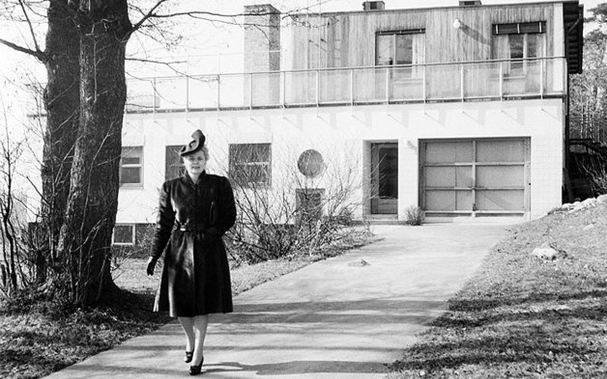 Svartvitt foto på Alva Myrdal. Iförd hatt går hon utanför huset i Bromma där hon bodde.