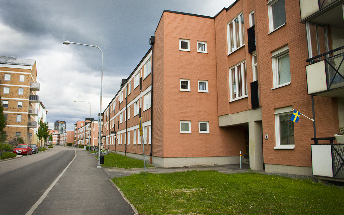 Uteplatser i Gränby. Gata med trevåningshus på höger sida med laxrosa fasader, vita detlajer och svart tak. Mörka moln på himlen.