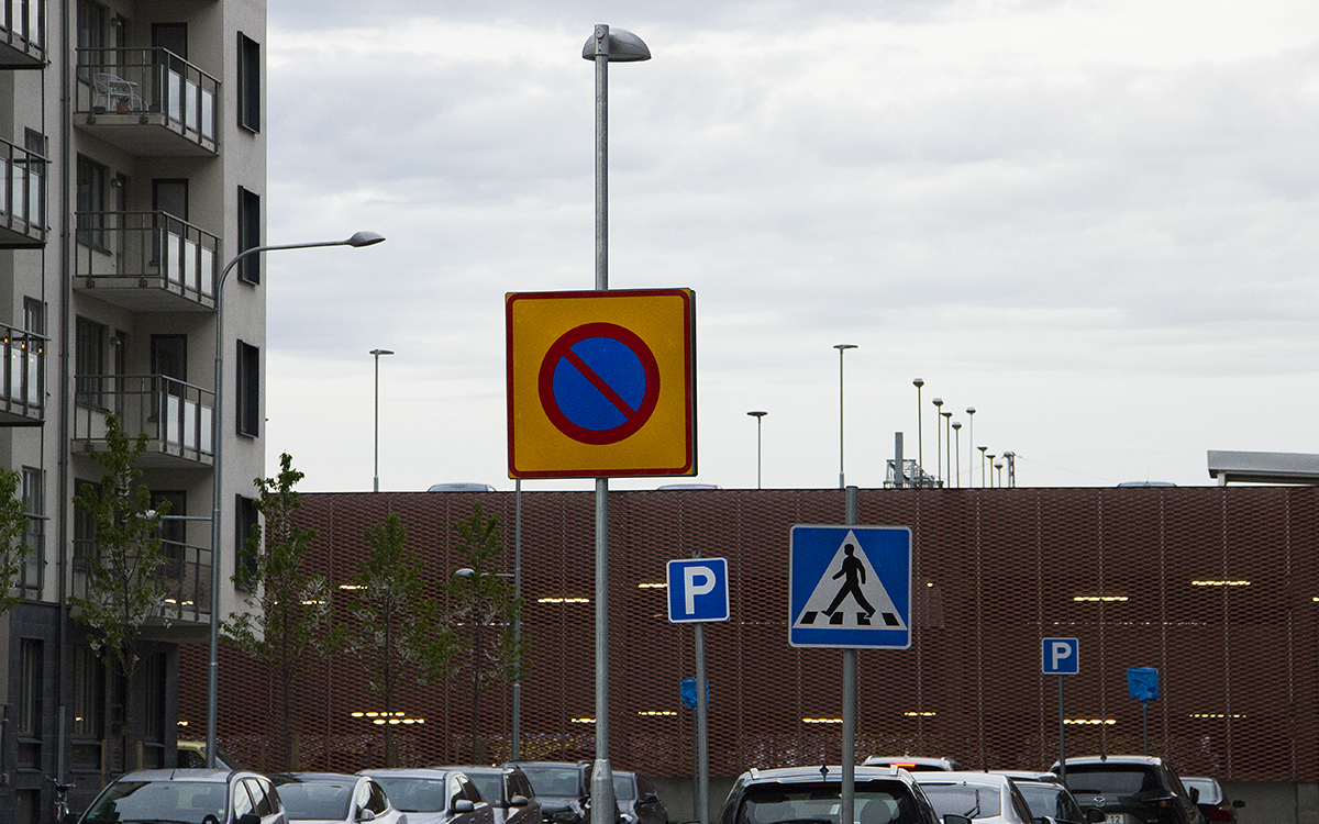 En områdesförbudsskylt och längre bak en översgångsställeskylt, två p-skyltar och två skyltar övertäckta av blå plast. Taken på många parkerade bilar.