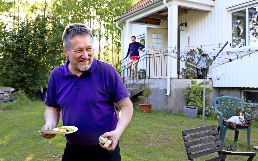 En man i lila tröja utanför ett vitt hus. På trappan en bit bort står en kvinna och ser ut över trädgården.