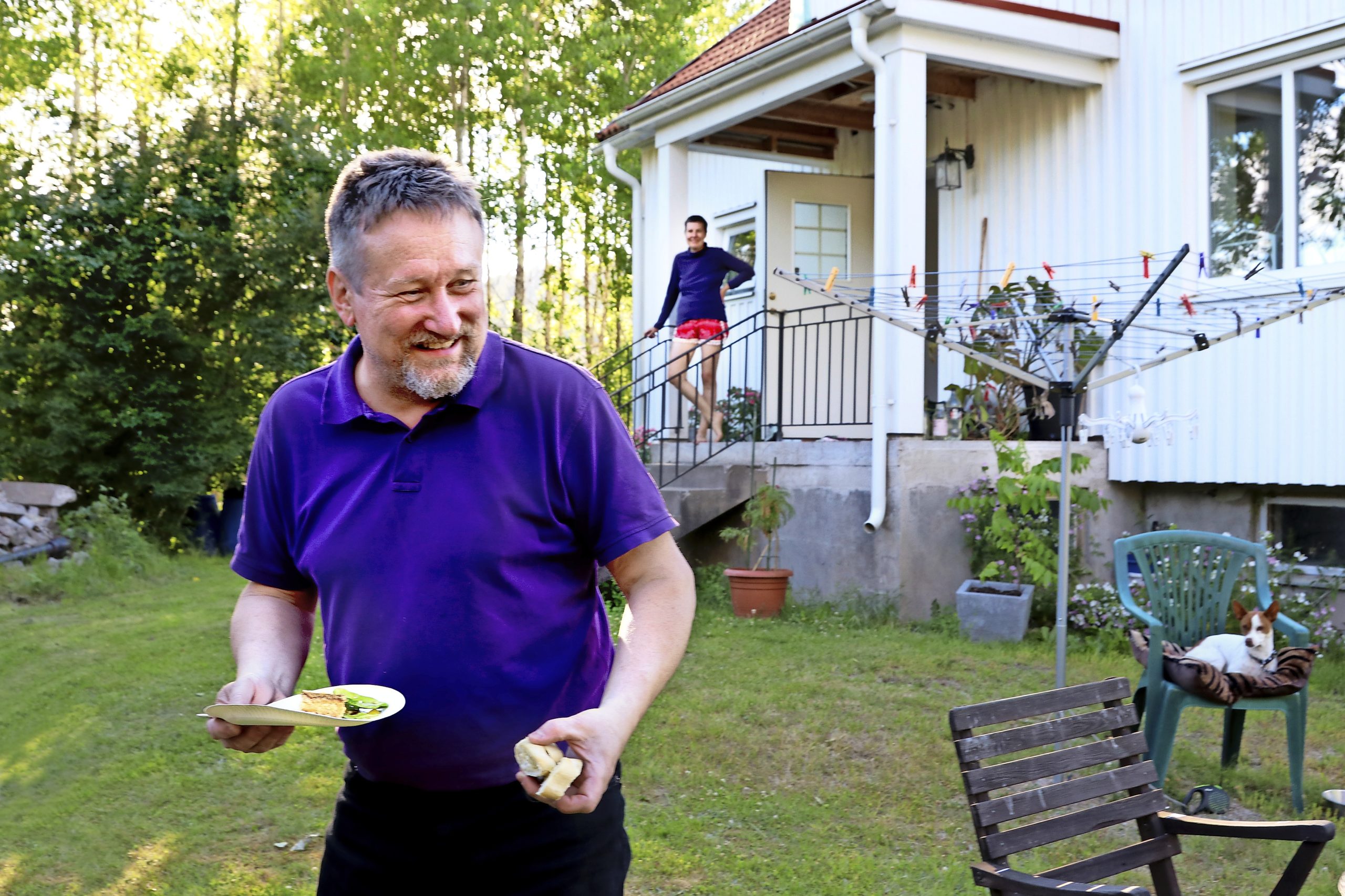 En man i lila tröja utanför ett vitt hus. På trappan en bit bort står en kvinna och ser ut över trädgården.