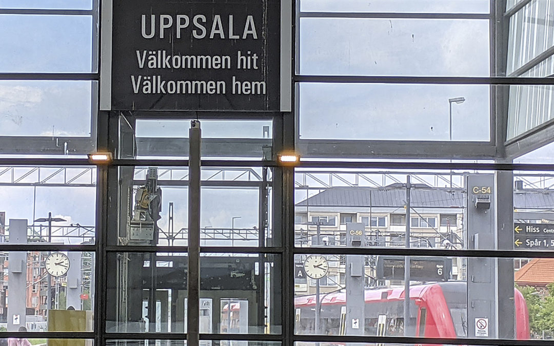 Skylt på Uppsala station. Uppsala kommun ger Bostad först en ny chans. UPPSALA Välkommen hit Välkommen hem. i Bakgrunden tågperrongen med ett tåg.