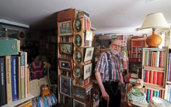 En äldre man står med käpp i ett rum fullt med böcker, möbler och tavlor.