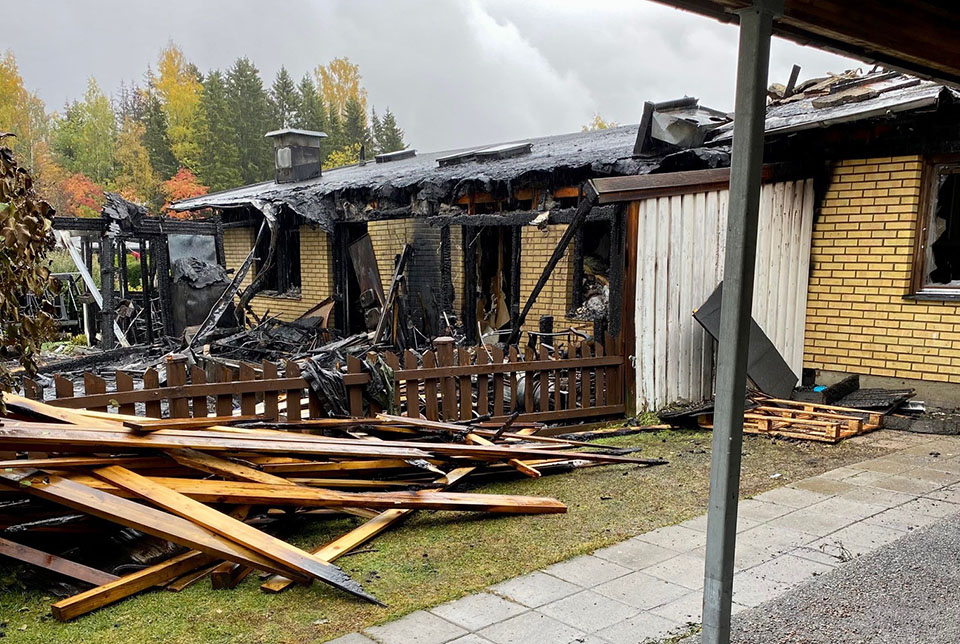 En eldhärjad radhuslänga med uppsamlat bråte efter branden på gräsmattan framför.