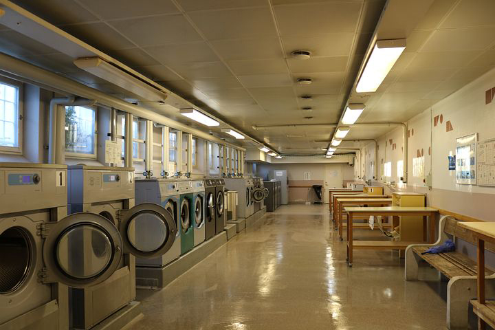 Bilden föreställer en tvättstuga i ljusrörsbelysning. Längs ena långväggen finns tvättmaskiner och ovan dem fönster. Längs andra långsidan finns sittbänkar, bord och manglar. 