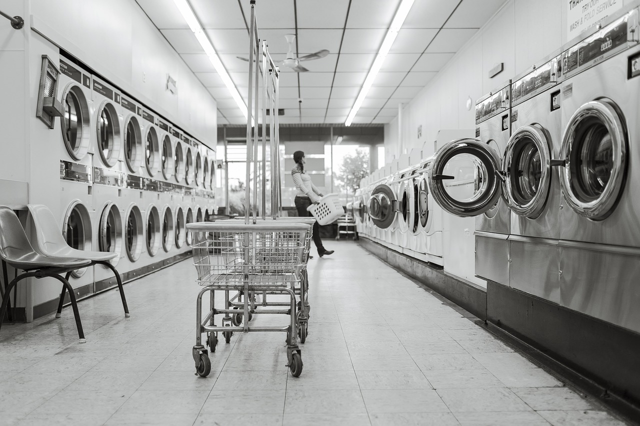 Bilden är i svartvitt och föreställer en tvättstuga. På båda sidor finns rader av tvättmaskiner. I mitten längst fram står ett par tvättvagnar. Längst bort finns et tfönster. Framför det syns en kvinna med en tvättkorg i händerna, redo att ladda en tvättmaskin med tvätt.