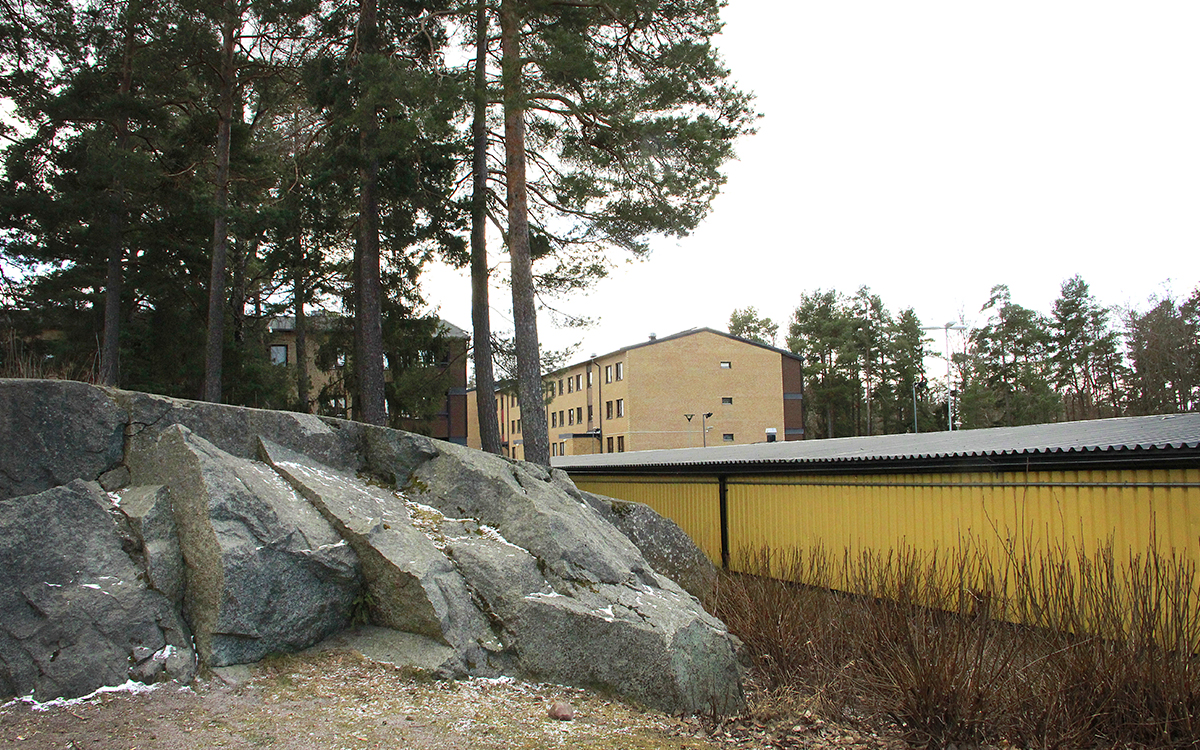 En berghäll, några tallar, baksidan av en gul garagelänga, frostigt gräs med lite snö i, längre bort trevåningshus i ljust tegel med bruna balkonger. Kvarter i Gottsunda.