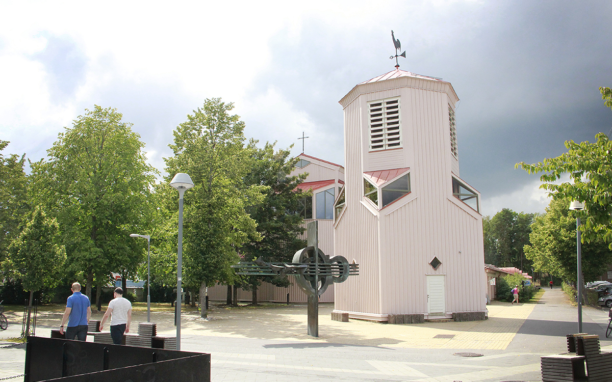 Gottsunda kyrka med klockstapeln i förgrunden, båda ljust laxrosa i trä med takpannor. Klockstpeln med en tupp i svart smide, kyrkobyggnaden med ett kors. Gröna träd och mörka moln på himlen, två personer i kortärmat går till vänster ur bilden. Framför kyrkan en skulptur.