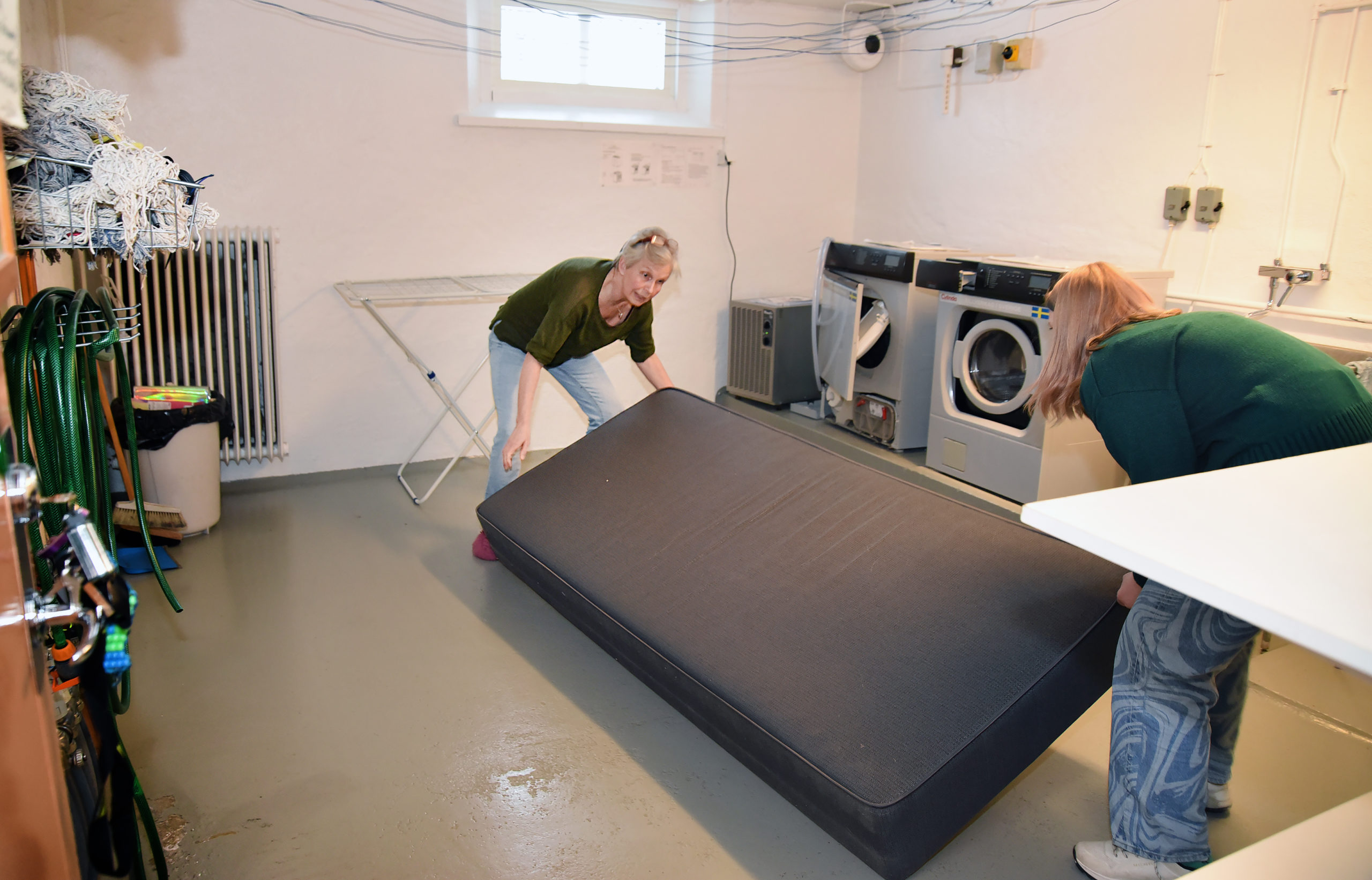 En 60-årig kvinna och hennes tonårsdotter hjälps åt att bära in en madrass i en tvättstuga.