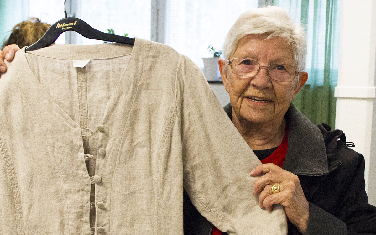 Anna Mikaelsson, halvkroppsporträtt där hon håller upp en klänning i ofärgat linne framför sig på en galge. Ana har vitt hår, glaögon, en röd tröja och en svart jacka med grå krage.