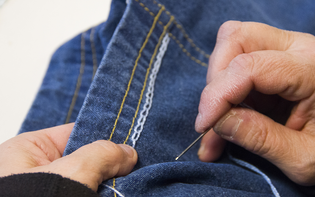 Närbild på jeanstyg och två händer med en nål i den högra handen, Tina Hägglund håller på att sy kedjesöm med ljusblå tråd.