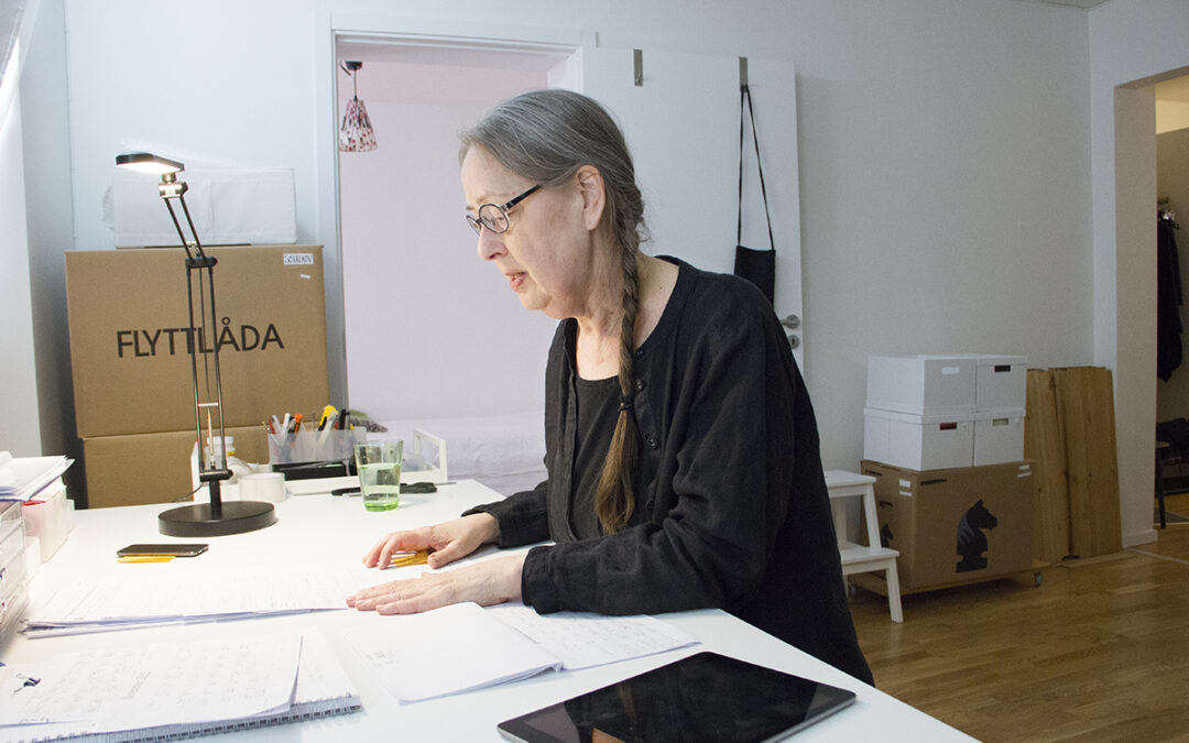 Kristina Lundgren, i svarta kläder, glasögon och långt hår i en fläta, sitter sedd i profil vid ett stort vitt skrivbord med papper, kalender i läsplatta. I bakgrunden syns flyttkartonger i hörnen. Flytten till seniorboendet blev en besvikelse.