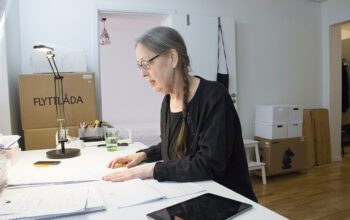 Kristina Lundgren, i svarta kläder, glasögon och långt hår i en fläta, sitter sedd i profil vid ett stort vitt skrivbord med papper, kalender i läsplatta. I bakgrunden syns flyttkartonger i hörnen. Flytten till seniorboendet blev en besvikelse.