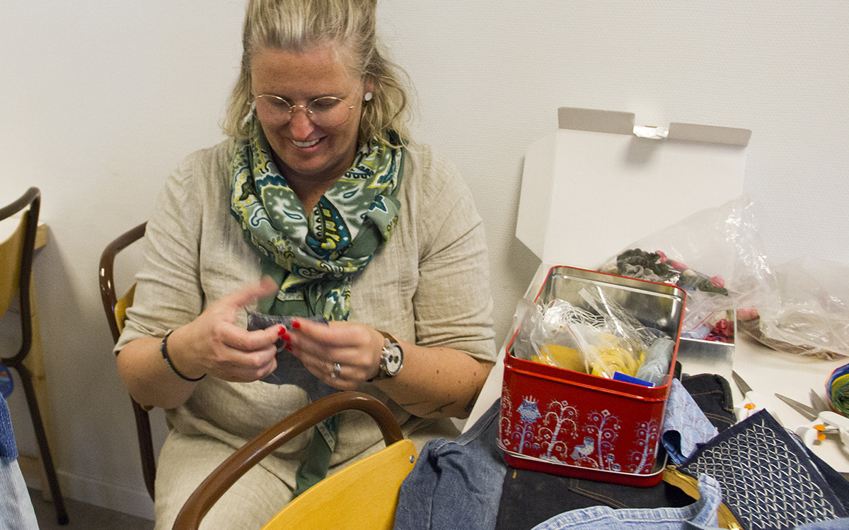 Maria Sjöndin, halvkroppsbild där hon sitter och broderar på en bit jeans. Maria har blont uppsatt hår, en grönmönstrad sjal runt halsen, runda glasögon. Till höger i bild ett bord med tyg och sybehör, en röd mönstrad plåtask, återbruksverkstad.