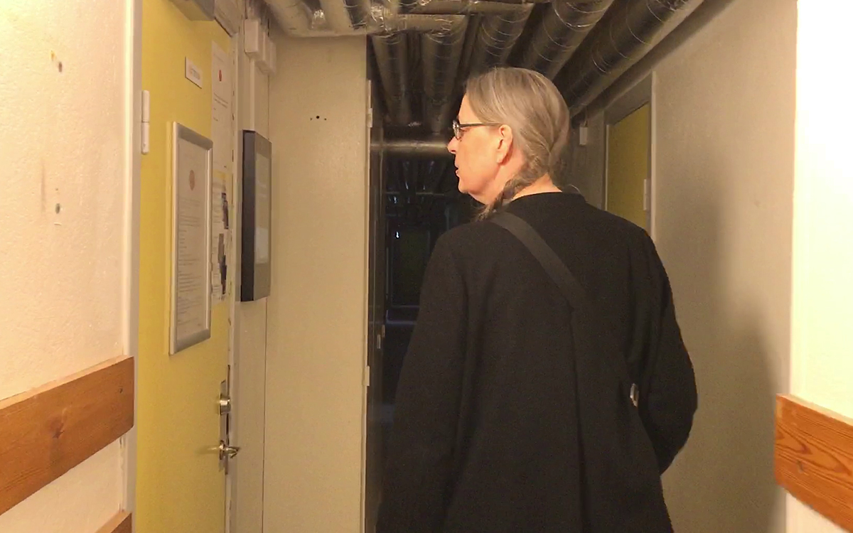 Kristina Lundgren, halvkroppsbild bakifrån, tittar till vänster på dörren till tvättstugan i en källarkorridor.