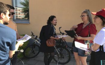 Åse Richard i röd kortärmad topp och jeansshorts, långt grått hår och glasögon, berättar ooch gestikulerar omgiven av deltagarna i stadsvandringen. De står intill en aprikosfärgad husvägg och i ett fönster speglas laxrosa bostadshus och grönska.