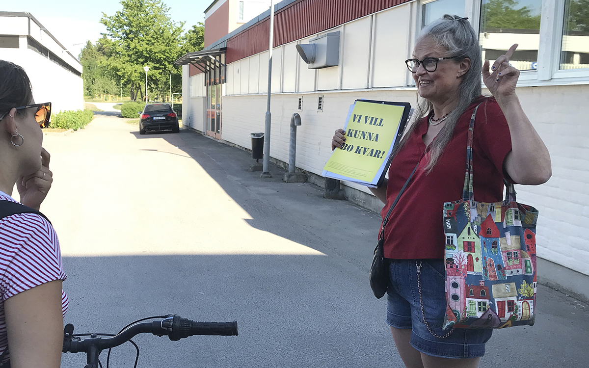 Åse Richard håller upp ett fult papper där det står "VI VILL KUNNA BO KVAR!" med ena handen, och har andra handen lyft med pekfingret i vädret. I vänster kant av bilden en av åhörarna bakifrån.