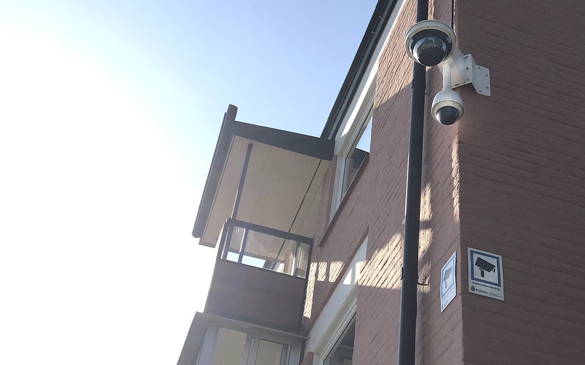 En övervakningskamera sitter högt upp på hörnet av ett laxrosa hus. Under den en skylt på varje husvägg, "Kamerabevakning", "Polisen".