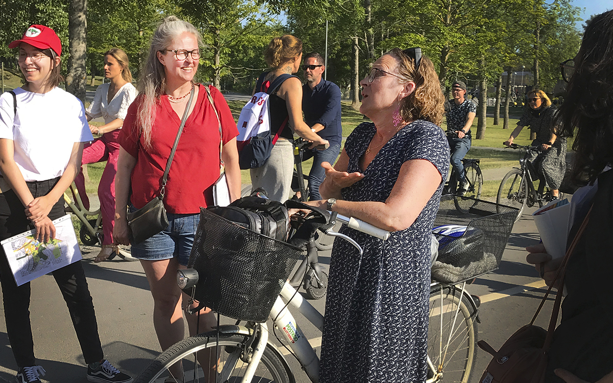 Bruna Teixeira, Åse Richard och Görel Forsling står på gångbanan när Görel pratar med deltagarna i stadsvandringen. Görel står med sin cykel med en väska i cykelkorgen. I bakgrunden en mängd cyklister och grönska.