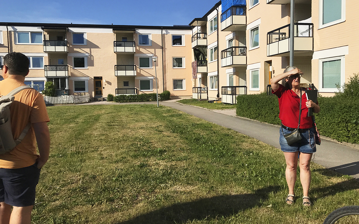 En innergård med enbart gräsmatta omgiven om laxrosa trevåningshus. Åse Richard står tll höger och skuggar ansiktet med en hand. Längst till vänster står Callum Ward med ryggen till och en ryggsäck på ryggen och tittar ut över innergården.