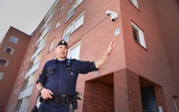 En polisklädd man, sedd lite underifrån, gestikulerar medan han talar. Bilden är tagen utomhus framför ett flerbostadshus. Uppe i ena hörnet syns en av polisens bevakningskameror.