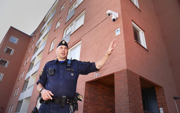 En polisklädd man, sedd lite underifrån, gestikulerar medan han talar. Bilden är tagen utomhus framför ett flerbostadshus. Uppe i ena hörnet syns en av polisens bevakningskameror.