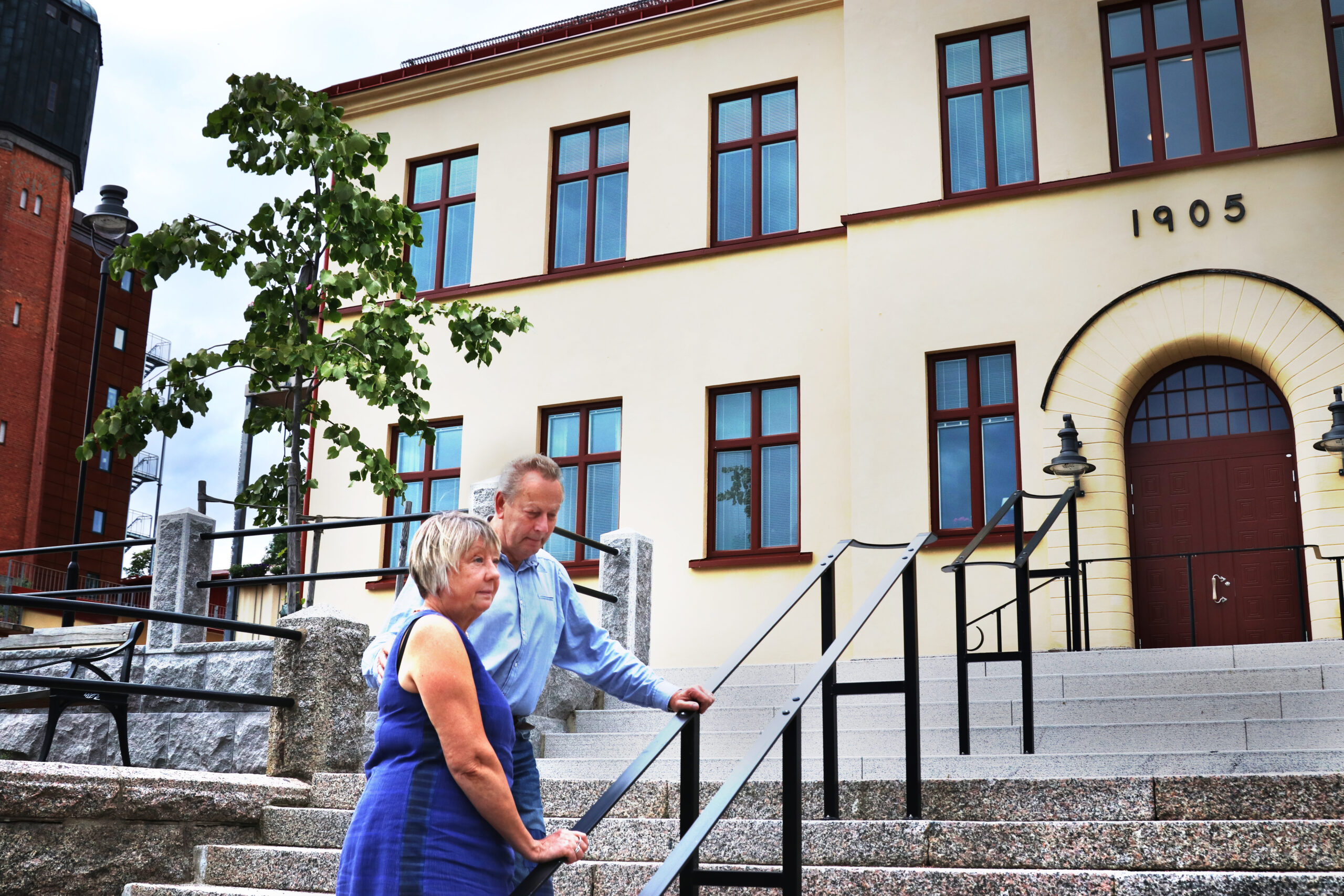 En man och en kvinna i blå kläder ståendes i en trappa utanför en gul byggnad.